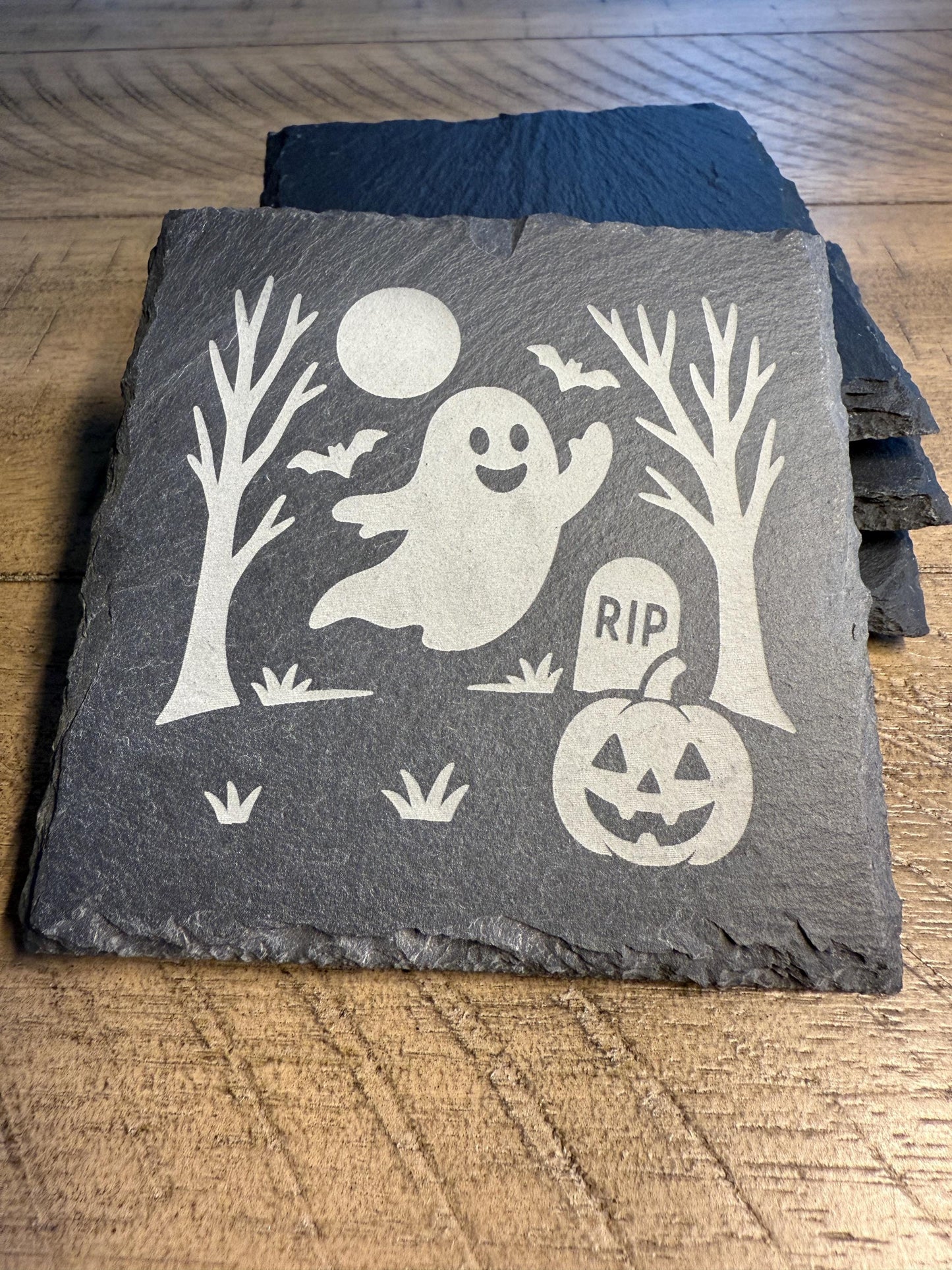 Slate coaster with Halloween design featuring a ghost, pumpkin, and tombstone on a wooden surface.