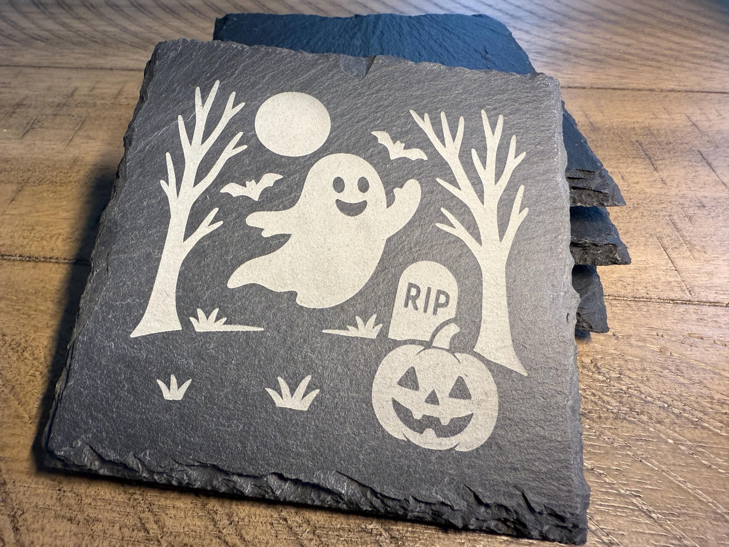 Slate coaster with Halloween design featuring a ghost, pumpkin, and tombstone on a wooden surface.