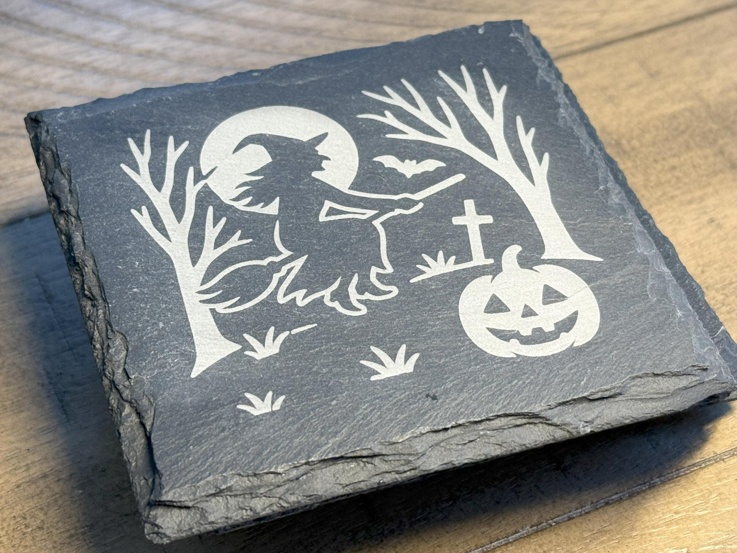 Slate coaster with Halloween design of a witch, moon, and pumpkin on a wooden surface