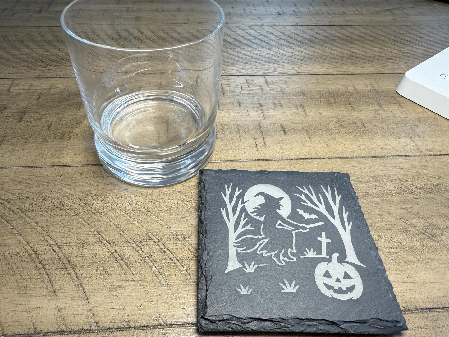 Slate coaster with Halloween design on a wooden surface next to a clear glass.