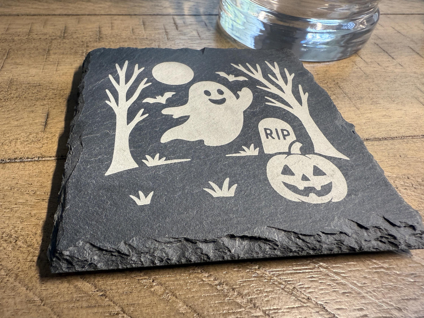 Slate coaster with Halloween design featuring a ghost, pumpkin, and tombstone on a wooden surface.