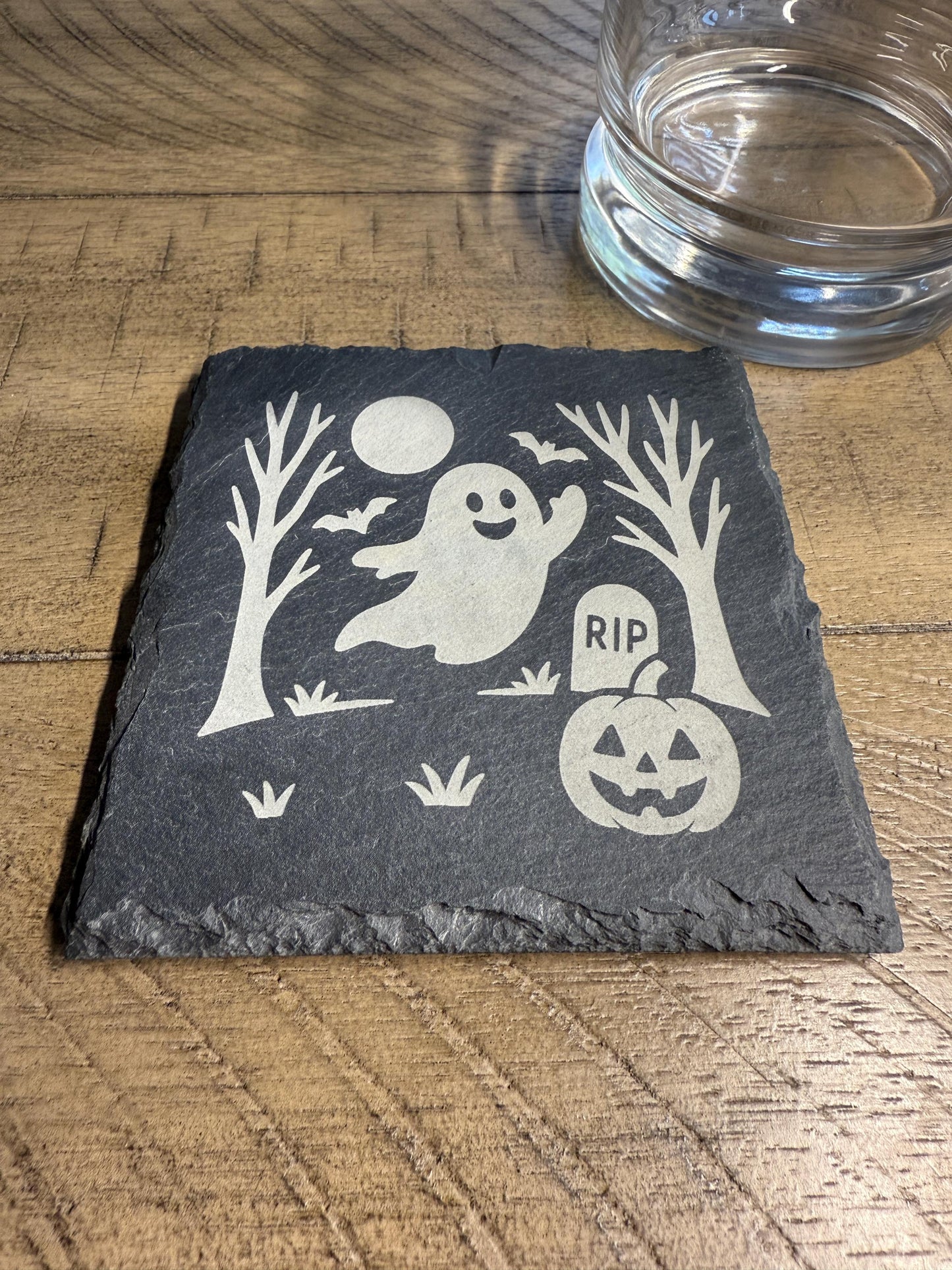 Slate coaster with Halloween design featuring a ghost, pumpkin, and tombstone on a wooden surface.
