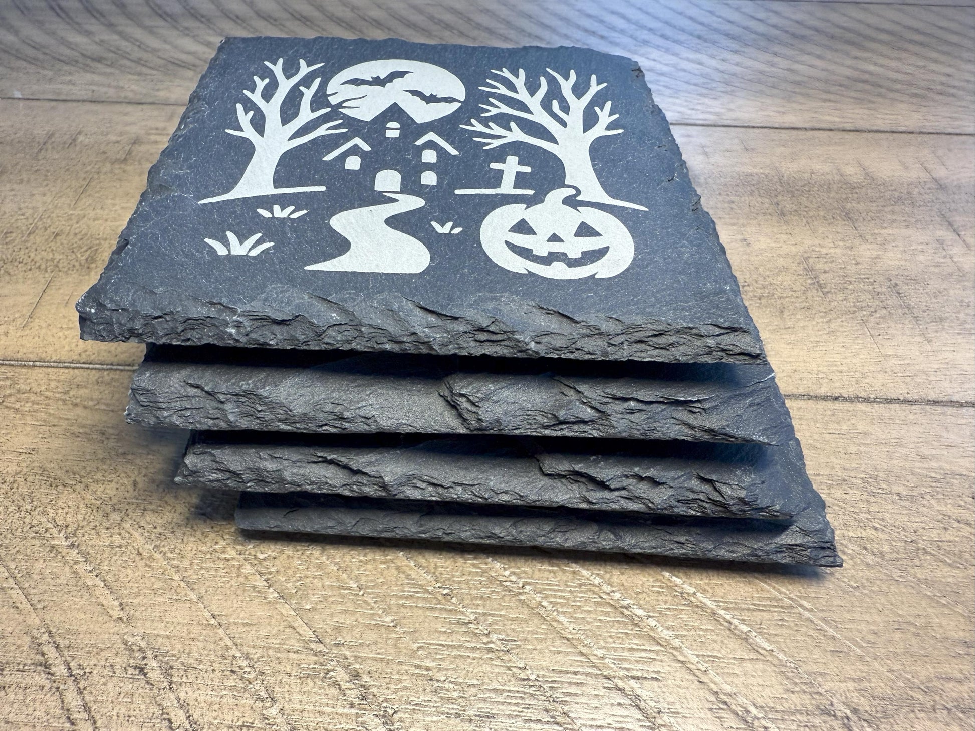 Set of four black slate coasters with Halloween-themed designs on a wooden surface.