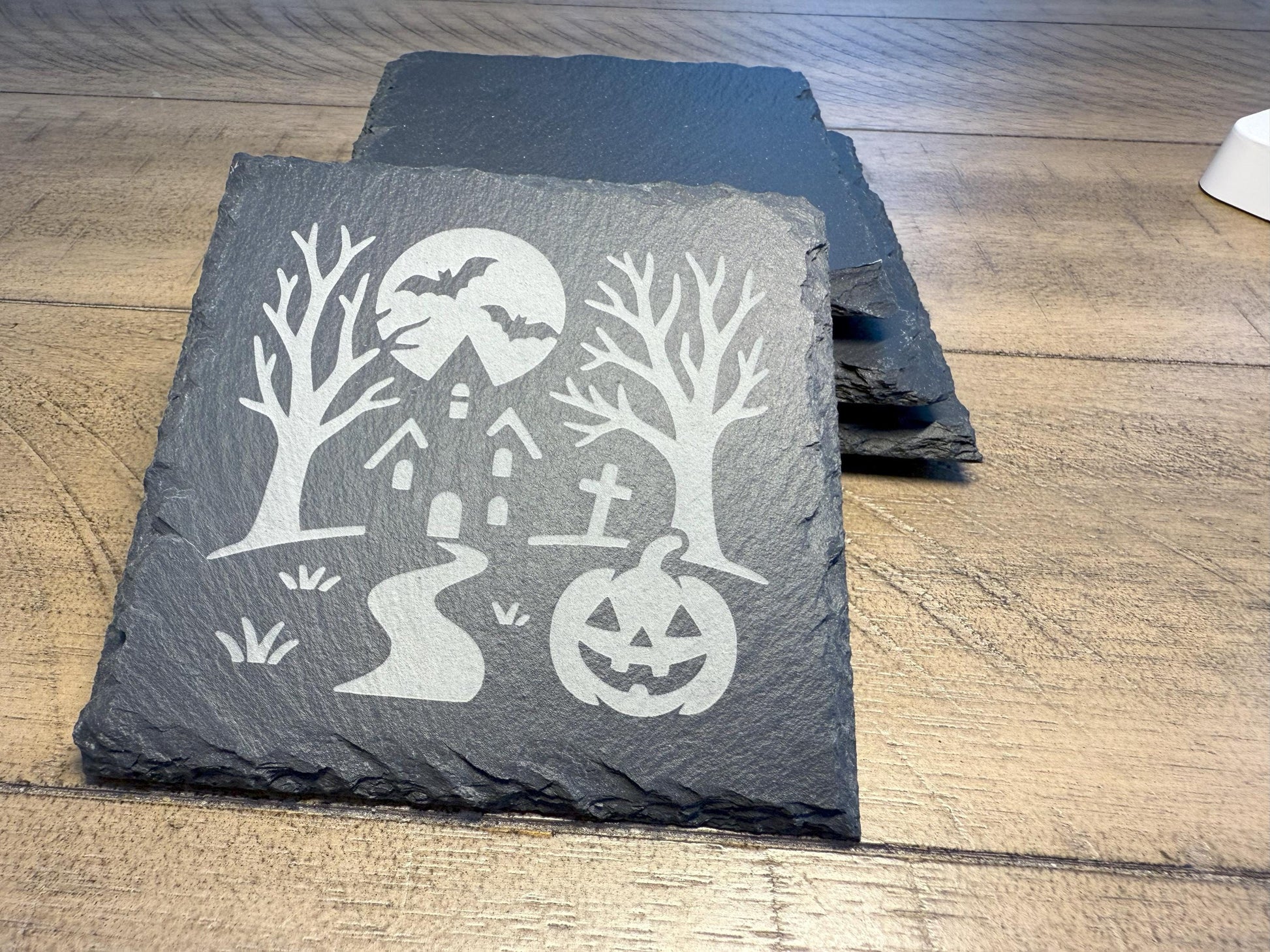 Slate coaster with Halloween design featuring a haunted house, trees, and pumpkins on a wooden surface.