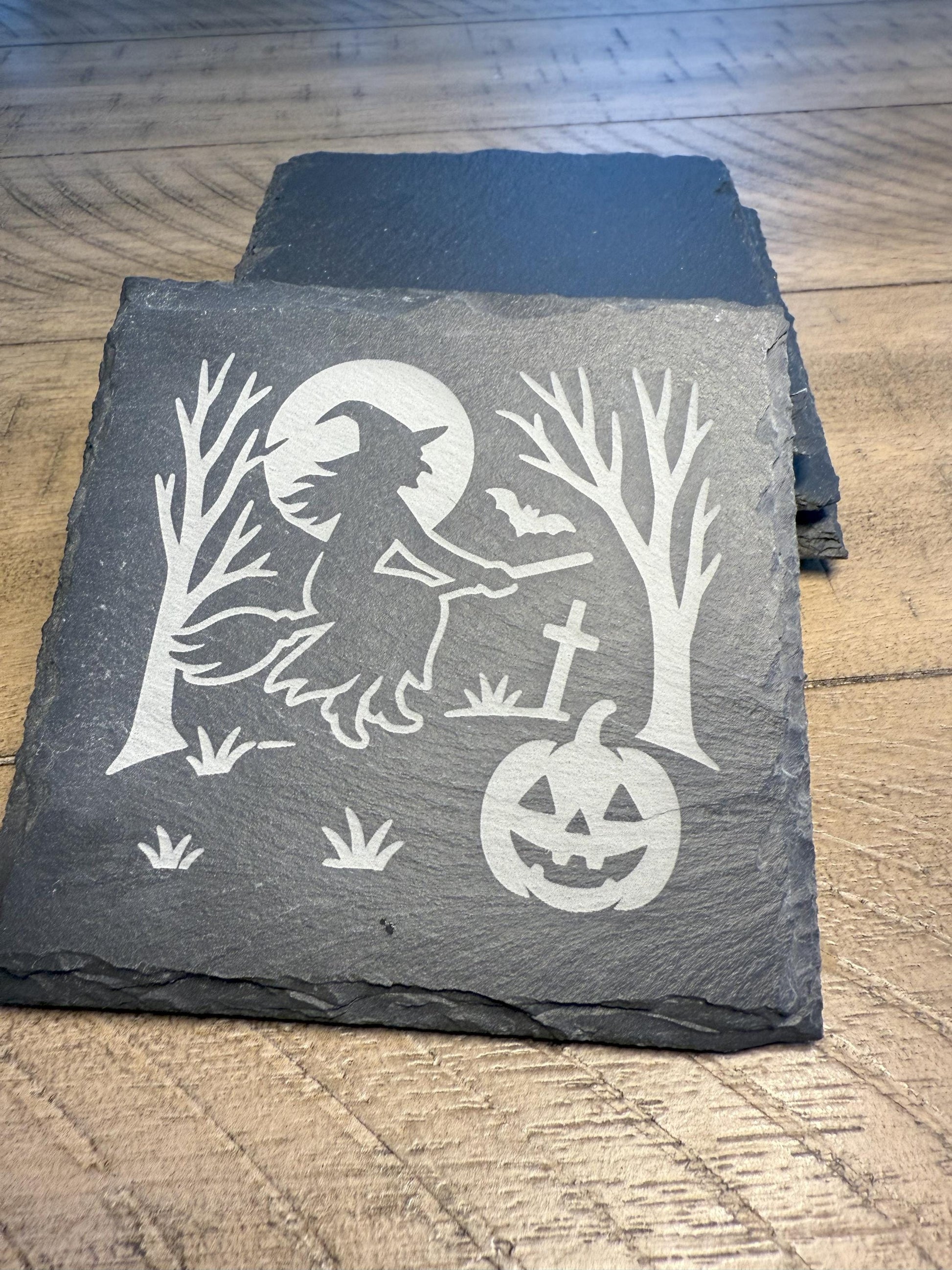 Slate coaster with Halloween design featuring a witch, pumpkin, and trees on a wooden surface.
