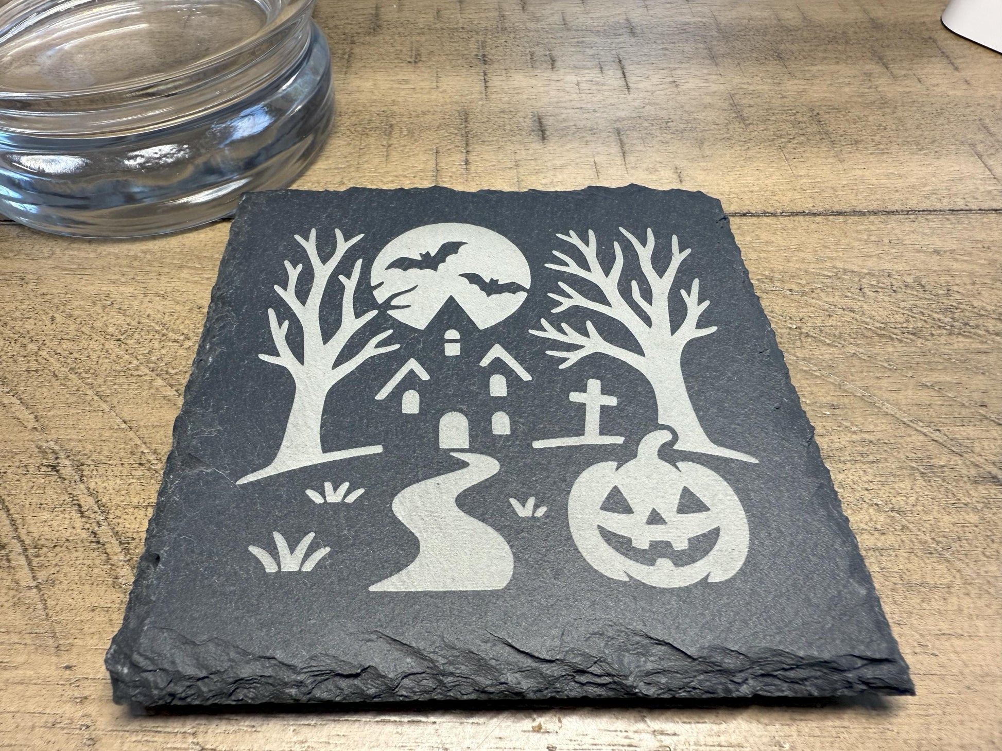 Slate coaster with Halloween design featuring a haunted house, jack-o'-lantern, and trees on a wooden surface.